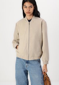 Beige pommikatel on ees lukuga, elastsete mansettidega ja küljetaskutega. Kombineeritud siniste teksade ja pruuni käekotiga. Sile tekstuur.