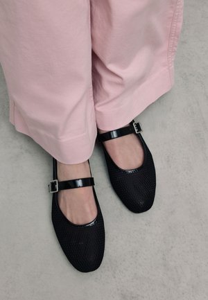 Black textured Mary Jane flats with glossy black straps and a silver buckle, paired with light pink wide-leg pants.