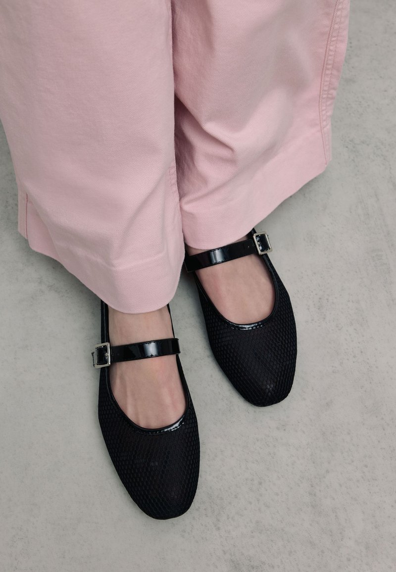 Zwarte, gestructureerde Mary Jane flats met glanzende zwarte banden en een zilveren gesp, gecombineerd met lichtroze wijde broek.