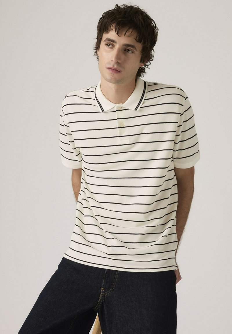 Joven con camiseta polo blanca con rayas horizontales negras y pantalones vaqueros oscuros, mirando ligeramente hacia un lado sobre un fondo liso.