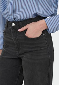 Jean en denim noir taille haute avec un design à cinq poches, fermeture à bouton argenté et détails de délavage léger, associé à un blouse à rayures bleues.