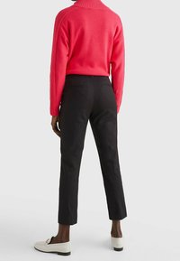 Person som bär svarta smala byxor, vita loafers och en röd ribbad höghalssweater, stående mot en enfärgad ljus bakgrund.