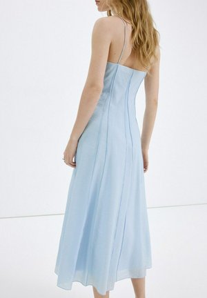 Kobieta o długich blond włosach, ubrana w jasnoniebieską sukienkę bez rękawów z cienkimi ramiączkami, zwrócona tyłem na białym tle.