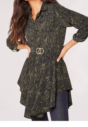 Femme portant une tunique à manches longues verte et noire à motifs, ceinturée d'une ceinture à double anneau, posant les mains sur les hanches devant un fond uni.