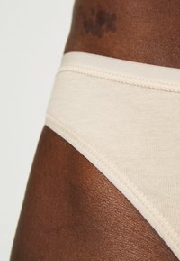 Beige Baumwollslip mit glatter Textur und genähten Kanten, teilweise vor dunkler Haut gezeigt. Einfaches Design ohne Muster oder Akzente.