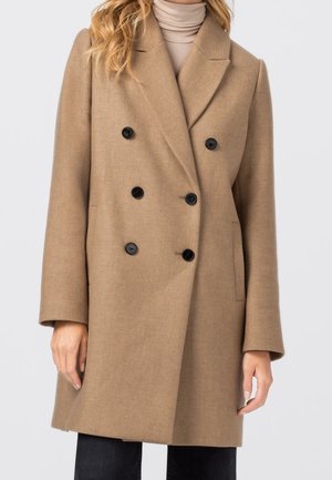 Dame camel-farvet dobbeltradet uldfrakke med sorte knapper, båret over beige rullekrave og sorte bukser.