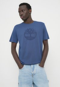 Μπλουζάκι από βαμβάκι σε ναυτικό μπλε χρώμα με γύρο λογότυπο δέντρου τυπωμένο σε ελαφρώς σκούρο μπλε στο στήθος. Κοντά μανίκια, στρογγυλή λαιμόκοψη, άνετη εφαρμογή.