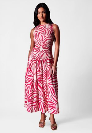 Femme aux longs cheveux foncés portant une robe midi sans manches rouge et blanche à motifs, ainsi que des talons ouverts marron, debout devant un fond uni.