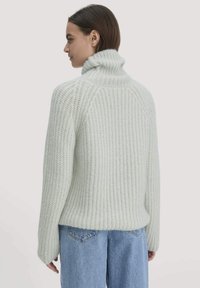Frau in einem hellgrauen, grob gestrickten Rollkragenpullover und blauen Jeans, die im Profil vor einem schlichten Hintergrund steht.