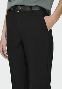 Pantaloni neri sartoriali con tasche laterali, realizzati in tessuto morbido, con taglio dritto e dettagli di cucitura sottili. Indossati con una cintura nera.