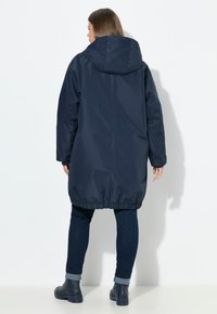 Mujer con cabello largo castaño, vistiendo un abrigo con capucha azul marino, jeans azul oscuro y botas negras, de pie contra un fondo blanco, vista desde atrás.