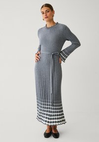 Graues, geripptes Strickkleid mit langen Ärmeln, tailliertem Gurt und gestreiftem Saum. Verfügt über einen Rüschenausschnitt und geschichtete Manschetten. Flache schwarze Schuhe.