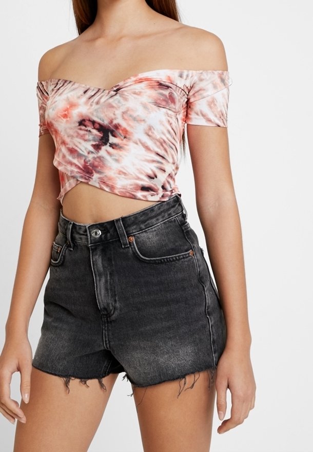 Off-the-shoulder, tie-dye kort topp i rosa och orange nyanser, parat med svarta högmidjade denimshorts med fransad nederkant.