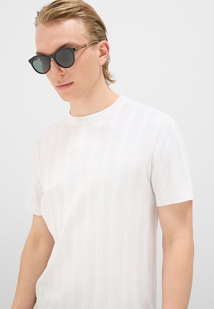 Giovane uomo con occhiali da sole neri e camicia bianca a maniche corte con sottili righe verticali, che guarda verso il basso e leggermente di lato.
