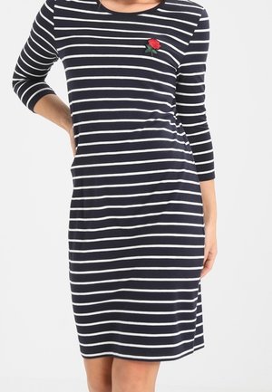 Kobieta ubrana w granatowo-białą, poziomo prążkowaną sukienkę do kolan z rękawami do połowy przedramienia oraz naszywką czerwonej róży na piersi.