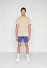 Camiseta deportiva beige de manga corta combinada con pantalones cortos azules con detalles en blanco. El modelo lleva zapatillas blancas y calcetines a media pierna.