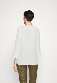 Donna con capelli corti e scuri, che indossa una blusa bianca a maniche lunghe e pantaloni verde oliva, girata di spalle, con un tatuaggio sul collo e orecchini d'oro.