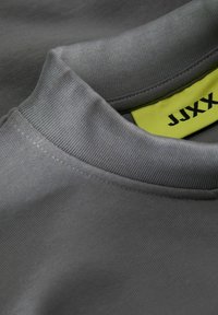 Šedé bavlněné tričko s žebrovaným kulatým výstřihem. Obsahuje jasně žlutou cedulku s textem "JJXX". Hladká textura s minimem švů.
