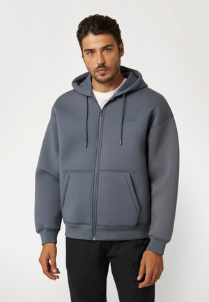 Pilkas zip-up džemperis su priekinėmis kišenėmis, reguliuojamu kapisonu ir logotipu. Pagamintas iš minkštos medžiagos ir laisvo kirpimo.