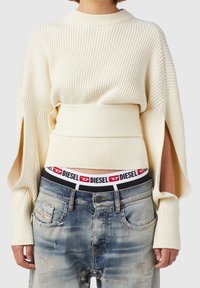 Suéter de crema acanalado con mangas amplias y cintura ceñida con cinturón, combinado con jeans azul desgastado que presentan un cinturón con el logo de Diesel.