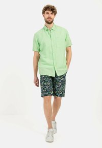 Helles grünes Kurzarmhemd mit einer Brusttasche, kombiniert mit marineblauen Shorts im bunten Blättermuster und weißen Sneakers.