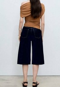 Brun off-shoulder top, mørkeblå vide denim culotter, set bagfra, med to baglommer med kontrastfarvet syning. Flade sandaler.
