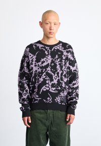 Pulover negru cu un model abstract mov, guler și manșete ribbate, fabricat din material tricotat, purtat cu pantaloni din catifea de culoare verde.