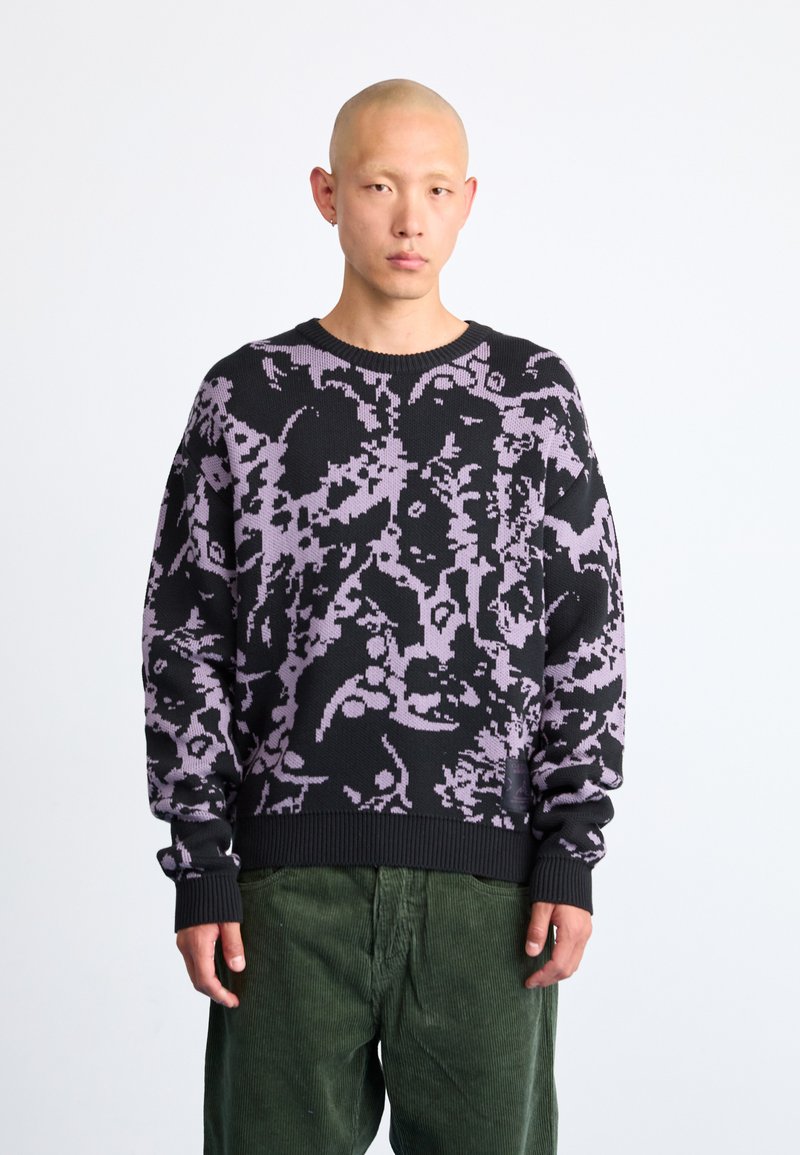 Pulover negru cu un model abstract mov, guler și manșete ribbate, fabricat din material tricotat, purtat cu pantaloni din catifea de culoare verde.