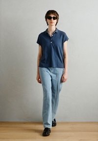 Námornícky modré polo tričko s krátkym rukávom a troj-button placket, skombinované s svetlomodrými džínami a čiernymi topánkami, nosené modelom vonku.