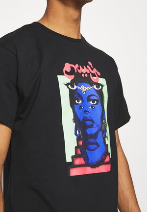 Personne portant un t-shirt noir avec un motif surréaliste multicolore représentant un visage bleu aux multiples yeux et des formes géométriques rouges.