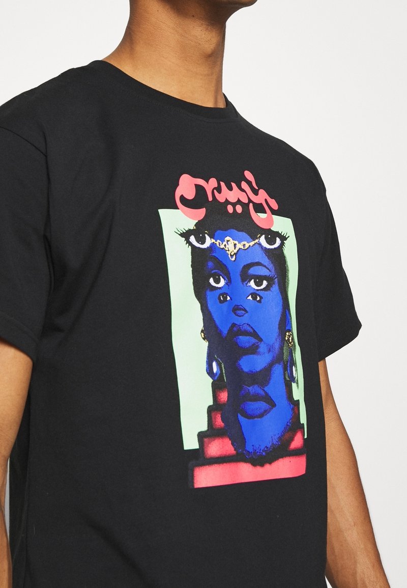 Personne portant un t-shirt noir avec un motif surréaliste multicolore représentant un visage bleu aux multiples yeux et des formes géométriques rouges.