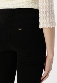 Jeans de mezclilla negra con forma ajustada, que cuentan con bolsillos traseros y una pequeña etiqueta amarilla con el logotipo "Lois". La tela parece suave con un ligero estiramiento.