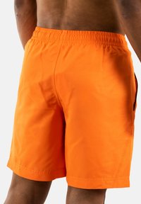 Vista posteriore di una persona che indossa pantaloni shorts atletici arancioni vivaci con una vita elasticizzata su uno sfondo semplice.