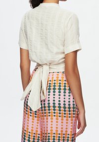 Blouse blanche texturée à manches courtes avec un lien à nouer dans le dos, associée à un pantalon rose orné de motifs à pois verts et oranges.