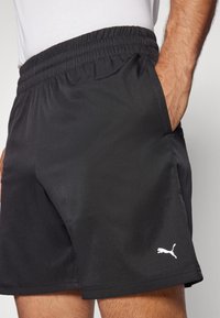 Hombre con pantalones cortos deportivos negros de Puma, con cinturilla elástica y mano en el bolsillo derecho, logo blanco de Puma en la pierna izquierda.