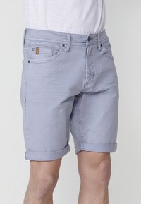 Gai zila denim šorti ar sarullētām malām, valkāti kopā ar baltu krekla, redzami priekšējie kabatas un jostas cilpas, uz gaiša fona.