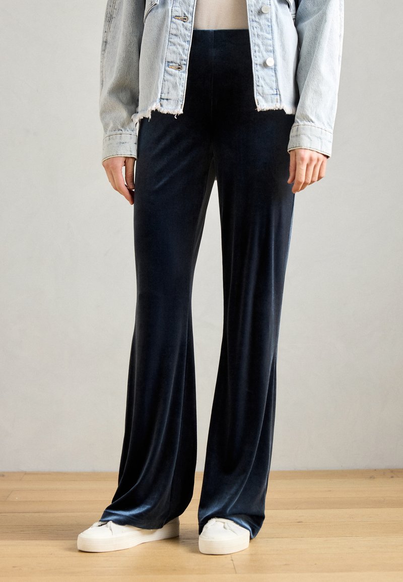 Jūras zila samta platā apakšdaļa ar gludu tekstūru, apvienota ar gaišu džinsa jaku un baltām auduma kedām uz koka grīdas.