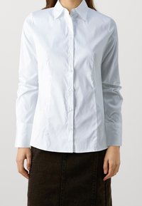 Camicia bianca con colletto strutturato, maniche lunghe e una texture liscia; abbinata a una gonna marrone scuro.