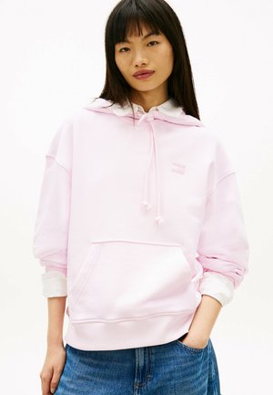 Jeune femme aux longs cheveux foncés portant un sweat à capuche rose clair par-dessus une chemise blanche, jean bleu, debout avec une main dans la poche.