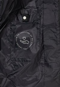 Innerseite einer schwarzen Nylonjacke mit einem Pflegeetikett, das eine Hand- und Herzgrafik zeigt. Enthält einen Druckknopf und gestickte Taschendetails.