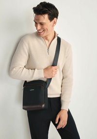 Czarna torba na ramię z teksturowanej tkaniny i brązowym skórzanym dnem, z zamknięciem na zamek błyskawiczny i regulowanym paskiem.