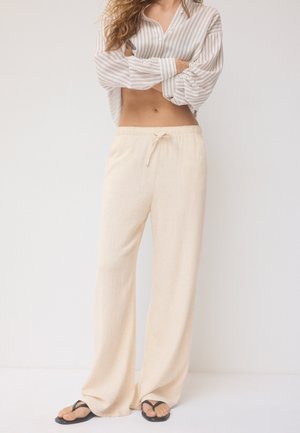 Kobieta ubrana w beżowe szerokie spodnie z troczkiem, pasiastą bluzkę z długim rękawem podniesioną do wysokości brzucha oraz czarne klapki, stojąca na jednolitym tle.