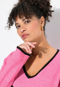 Rosa tröja med V-neck och svart kant. Mjuk textur. Modellen poserar med handen på hakan, bär stora guldörhängen. Bakgrunden är vit.