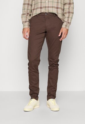 Mann trägt braune, schmal geschnittene Hose, beige kariertes Langarmhemd und cremeweiße Sneaker, steht vor einem schlichten Hintergrund.