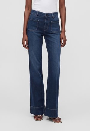 Kvinde iført mørkeblå højtaljede vidde jeans med forlommer og opbukket kant, kombineret med en hvid top og sandaler.