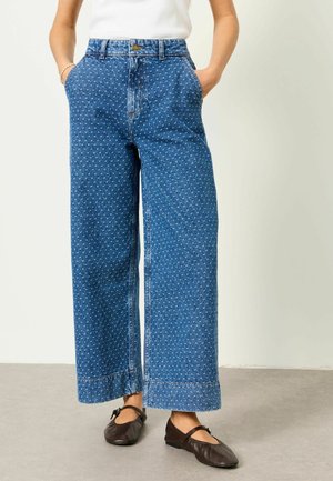 Naine kannab kõrge vöökohaga laia jala siniseid teksapükse, millel on valged väikesed lillemustrid, valget topi ja tumedat pruunikat Mary Jane stiilis kingi, käed taskutes.