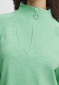 Pull vert menthe avec un col montant à texture côtelée, une fermeture éclair frontale et une coupe ajustée avec des détails de surpiqûres.