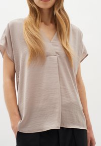 Blusa corta a maniche corte di un colore taupe chiaro con scollo a V e tessuto morbido e testurizzato. Presenta una vestibilità ampia e sottili pieghe davanti.