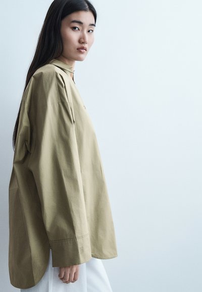 Jeune femme aux longs cheveux foncés portant une chemise beige surdimensionnée, debout de profil sur un fond clair uni.