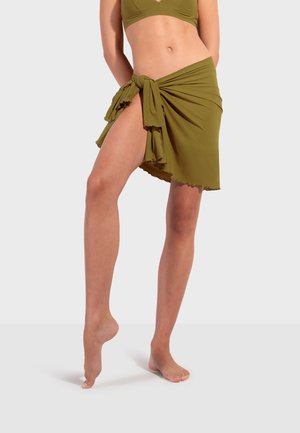 Žena v olivovo zelenom bikinách a s dobieraným sarongom zaviazaným na bok, stojaca na špičke jednej nohy na jednoduchom pozadí.